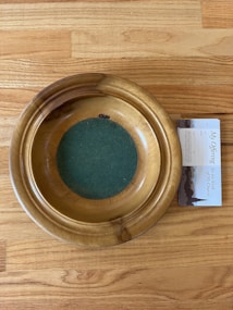 A wooden offering plate with a green felt base is placed on a light wooden floor. Next to the plate, there is a pamphlet partially visible, featuring text and images related to church offerings.