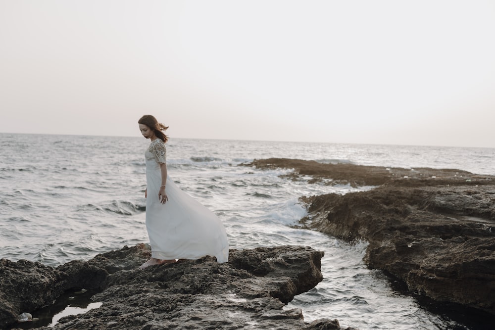 uma pessoa em um vestido branco de pé em uma praia rochosa