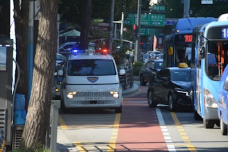 A busy urban street with a police vehicle using its lights, navigating through traffic. Several buses and cars are visible on the road, along with trees and traffic signs.