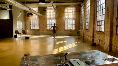 A spacious dance studio with large windows allowing plenty of natural light to pour in, casting shadows on the yellow floor. A lone dancer is practicing near the barre in the background. The room has exposed brick walls and industrial elements, including a large ventilation duct on the ceiling. Near the foreground, there is a table with signs, 'World's Best Student,' notebooks, a microphone, and several glasses. A few chairs are positioned to the side.