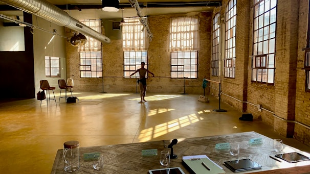 A spacious dance studio with large windows allowing plenty of natural light to pour in, casting shadows on the yellow floor. A lone dancer is practicing near the barre in the background. The room has exposed brick walls and industrial elements, including a large ventilation duct on the ceiling. Near the foreground, there is a table with signs, 'World's Best Student,' notebooks, a microphone, and several glasses. A few chairs are positioned to the side.