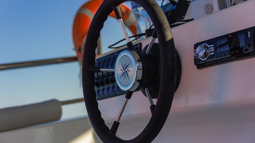 A close-up view of a boat's steering wheel with a nautical star design at the center. The dashboard features various controls and a stereo system. In the background, a lifebuoy is visible, secured by metal railings. The lighting suggests it's either morning or late afternoon with a clear sky.