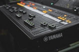 A close-up view of a Yamaha audio mixing console with multiple sliders and illuminated buttons. The surface features various dials and control switches, predominantly in a dark color scheme with green and orange lights creating a dynamic visual contrast.