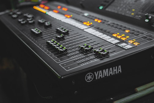 A close-up view of a Yamaha audio mixing console with multiple sliders and illuminated buttons. The surface features various dials and control switches, predominantly in a dark color scheme with green and orange lights creating a dynamic visual contrast.