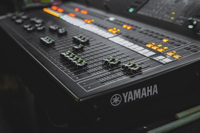 A close-up view of a Yamaha audio mixing console with multiple sliders and illuminated buttons. The surface features various dials and control switches, predominantly in a dark color scheme with green and orange lights creating a dynamic visual contrast.