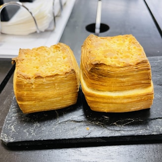 Two square pastries with multiple flaky layers are placed on a dark slate surface in a kitchen setting. The pastries have a golden brown crust with visible layers, resembling croissants or puff pastries.