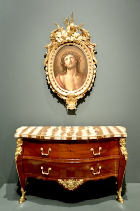 An ornate framed painting featuring a person with a crown of thorns is mounted above a luxurious wooden dresser. The dresser has a polished finish and features gold accents, including intricate handles and decorative details on its legs and edges.