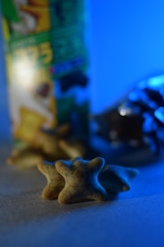 Three star-shaped snacks with intricate patterns are in focus, set against a blurred background of a snack box and shiny foil packaging. The scene is illuminated with a cool blue light, creating a striking contrast against the snacks' pale colors.