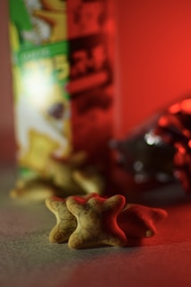 A close-up of star-shaped snacks with a detailed pattern on them, placed in front of a colorful snack package and a foil-wrapped item. The lighting has a warm, reddish tone.