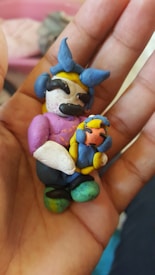 A hand holding a small, handmade clay figure. The figure appears to be a cartoonish character with exaggerated features, wearing a blue and yellow headdress with a bow, and holding a smaller figure.