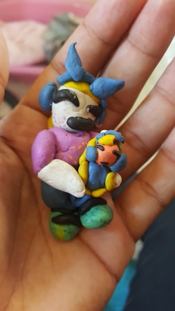 A hand holding a small, handmade clay figure. The figure appears to be a cartoonish character with exaggerated features, wearing a blue and yellow headdress with a bow, and holding a smaller figure.