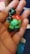 A small, handmade figurine made from colorful modeling clay, featuring a mix of green, red, and black colors. The figure appears to have a humanoid shape with exaggerated features, and is being held in the palm of a hand.