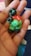 A small, handmade figurine made from colorful modeling clay, featuring a mix of green, red, and black colors. The figure appears to have a humanoid shape with exaggerated features, and is being held in the palm of a hand.