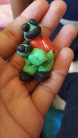A small, handmade figurine made from colorful modeling clay, featuring a mix of green, red, and black colors. The figure appears to have a humanoid shape with exaggerated features, and is being held in the palm of a hand.
