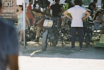 Several people are gathered outdoors, possibly at a motorcycle event or race. A motorcycle with the number 24 is prominently positioned in the center, with various attendees standing and conversing nearby. There are chairs and tents around, suggesting a casual, social gathering. The atmosphere seems informal and lively.