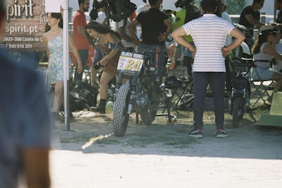 Several people are gathered outdoors, possibly at a motorcycle event or race. A motorcycle with the number 24 is prominently positioned in the center, with various attendees standing and conversing nearby. There are chairs and tents around, suggesting a casual, social gathering. The atmosphere seems informal and lively.