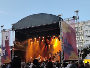 An outdoor concert stage is set up with a large canopy overhead. Bright stage lights in yellow and orange illuminate the performers, creating a vibrant atmosphere. A crowd of people stands in front, watching the musicians on stage. The backdrop displays abstract art and buildings are visible in the background under a clear sky.