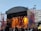 An outdoor concert stage is set up with a large canopy overhead. Bright stage lights in yellow and orange illuminate the performers, creating a vibrant atmosphere. A crowd of people stands in front, watching the musicians on stage. The backdrop displays abstract art and buildings are visible in the background under a clear sky.
