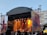 An outdoor concert stage is set up with a large canopy overhead. Bright stage lights in yellow and orange illuminate the performers, creating a vibrant atmosphere. A crowd of people stands in front, watching the musicians on stage. The backdrop displays abstract art and buildings are visible in the background under a clear sky.