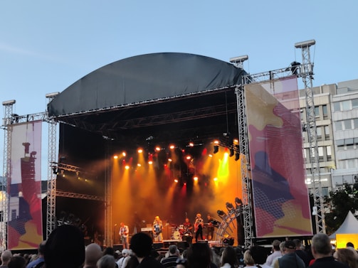 An outdoor concert stage is set up with a large canopy overhead. Bright stage lights in yellow and orange illuminate the performers, creating a vibrant atmosphere. A crowd of people stands in front, watching the musicians on stage. The backdrop displays abstract art and buildings are visible in the background under a clear sky.