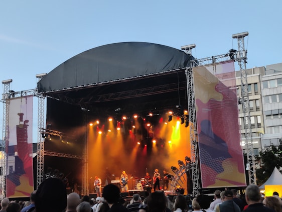 An outdoor concert stage is set up with a large canopy overhead. Bright stage lights in yellow and orange illuminate the performers, creating a vibrant atmosphere. A crowd of people stands in front, watching the musicians on stage. The backdrop displays abstract art and buildings are visible in the background under a clear sky.