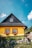 A charming wooden house with a vibrant yellow exterior and a black roof. The windows are framed with red trim and adorned with flower boxes, overflowing with greenery and flowers. The house sits amidst lush grass and stones, under a bright blue sky with a few clouds.
