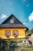 A charming wooden house with a vibrant yellow exterior and a black roof. The windows are framed with red trim and adorned with flower boxes, overflowing with greenery and flowers. The house sits amidst lush grass and stones, under a bright blue sky with a few clouds.