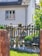 A garden scene with a rustic wooden fence adorned with potted plants. A statue of an elderly man holding a succulent is positioned near a bench surrounded by decorative objects and stones. Lush greenery and a quaint house with a tiled roof provide a serene, homey backdrop.