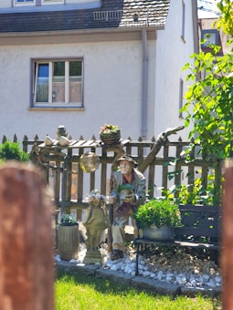 A garden scene with a rustic wooden fence adorned with potted plants. A statue of an elderly man holding a succulent is positioned near a bench surrounded by decorative objects and stones. Lush greenery and a quaint house with a tiled roof provide a serene, homey backdrop.