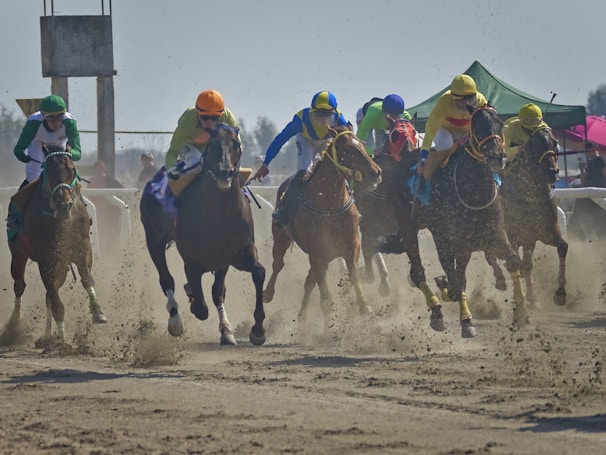 A group of jockeys in colorful uniforms and helmets are riding horses at full speed on a dirt racetrack. The horses are kicking up dust as they race forward, surrounded by a cloud of dust. In the background, a structure and tents are visible under a clear sky.