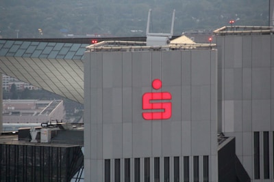 A large, modern building with a prominent red logo of a financial institution on its façade. The building has a geometric and industrial design, surrounded by other urban structures. A rooftop with various equipment and antennas can be seen, along with a cityscape in the background.
