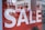 Large white letters spelling 'SALE' are displayed on a bright red background. The text is prominently positioned, and in the lower part of the image, a glimpse of a shoe is visible inside a store.