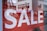 Large white letters spelling 'SALE' are displayed on a bright red background. The text is prominently positioned, and in the lower part of the image, a glimpse of a shoe is visible inside a store.