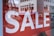 Large white letters spelling 'SALE' are displayed on a bright red background. The text is prominently positioned, and in the lower part of the image, a glimpse of a shoe is visible inside a store.