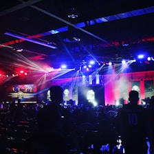 A dimly lit indoor arena filled with an audience. Bright stage lights in red and blue illuminate multiple large screens displaying graphics.