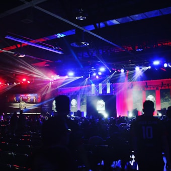 A dimly lit indoor arena filled with an audience. Bright stage lights in red and blue illuminate multiple large screens displaying graphics.