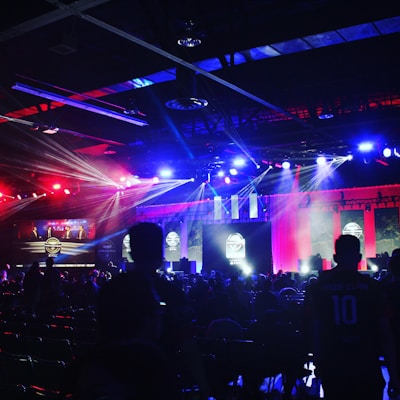 A dimly lit indoor arena filled with an audience. Bright stage lights in red and blue illuminate multiple large screens displaying graphics.