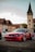 A red Lexus car is parked on a cobbled street with historic buildings in the background. The sky is partly cloudy, and a prominent clock tower is visible, adding a touch of classic architecture to the scene.