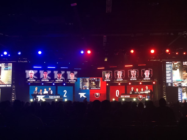 An esports event with a large screen displaying two teams, Team Kaliber and Rise Nation, competing against each other. The screen shows profiles of team members and scores, with Team Kaliber leading 2-0. The venue is dimly lit with blue and red lighting, and an audience can be seen watching.