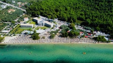 An aerial view of a beachfront area with a densely forested region in the background. There are several buildings, possibly hotels or resorts, alongside a busy road. The beach is lined with numerous umbrellas and sun loungers, and many people are visible enjoying the water and sand. The shoreline meets clear turquoise water, with a few swimmers and a small yellow inflatable object visible in the sea.