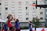 A group of young people are playing basketball outside. One player in a red jersey is jumping to shoot the ball towards the hoop, while two other players in blue jerseys are attempting to block or defend. The game takes place in an urban setting with a large residential building in the background. The sky appears clear, suggesting it's an outdoor court, possibly in a recreational area.