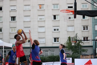 A group of young people are playing basketball outside. One player in a red jersey is jumping to shoot the ball towards the hoop, while two other players in blue jerseys are attempting to block or defend. The game takes place in an urban setting with a large residential building in the background. The sky appears clear, suggesting it's an outdoor court, possibly in a recreational area.