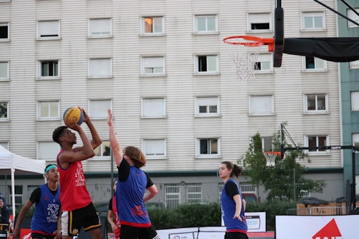 A group of young people are playing basketball outside. One player in a red jersey is jumping to shoot the ball towards the hoop, while two other players in blue jerseys are attempting to block or defend. The game takes place in an urban setting with a large residential building in the background. The sky appears clear, suggesting it's an outdoor court, possibly in a recreational area.