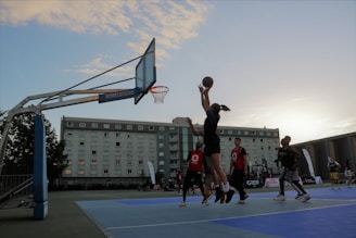 A group of young people are actively playing basketball on an outdoor court during sunset. One player is jumping with a basketball aiming for the hoop, while others watch or prepare to defend. The background features a residential building and a few spectators.