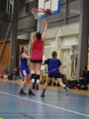 A group of female basketball players are actively participating in a game inside a sports hall. One player, wearing a red jersey, is jumping and reaching towards the basketball hoop with the ball. Two other players in blue jerseys are nearby, attempting to either defend or block the play. In the background, there are spectators or team members sitting and watching the game.