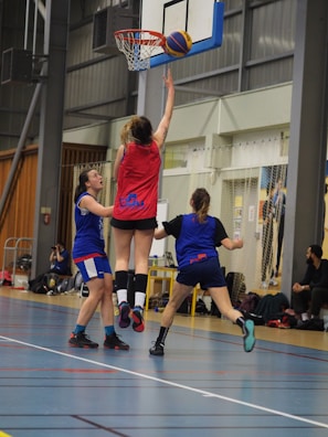 A group of female basketball players are actively participating in a game inside a sports hall. One player, wearing a red jersey, is jumping and reaching towards the basketball hoop with the ball. Two other players in blue jerseys are nearby, attempting to either defend or block the play. In the background, there are spectators or team members sitting and watching the game.