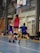 A group of female basketball players are actively participating in a game inside a sports hall. One player, wearing a red jersey, is jumping and reaching towards the basketball hoop with the ball. Two other players in blue jerseys are nearby, attempting to either defend or block the play. In the background, there are spectators or team members sitting and watching the game.