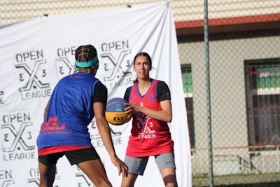 Two basketball players on an outdoor court engaged in a game. They are wearing jerseys with logos and appear focused on the action. A branded event backdrop and a chain-link fence are visible in the background.