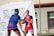 Two basketball players on an outdoor court engaged in a game. They are wearing jerseys with logos and appear focused on the action. A branded event backdrop and a chain-link fence are visible in the background.