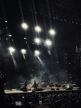 A concert scene with a band performing on stage under a series of bright spotlights. Smoke effects enhance the atmosphere as the audience watches. Musical instruments like guitars, keyboards, and drums are visible, and the performers are silhouetted against the illuminated background.